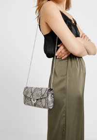 Kabelka s hadím vzorem v šedé a černé barvě, se skládacím uzávěrem a stříbrným řetízkem. Hladký povrch s lesklým povrchem.