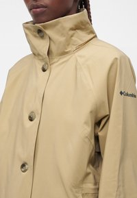 Gros plan d’une personne portant une veste imperméable beige boutonnée avec un col montant et le logo Columbia sur la manche gauche.