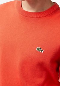 Zbliżenie osoby w jasnopomarańczowym swetrze z okrągłym dekoltem, z małym zielonym logo krokodyla na lewej piersi.