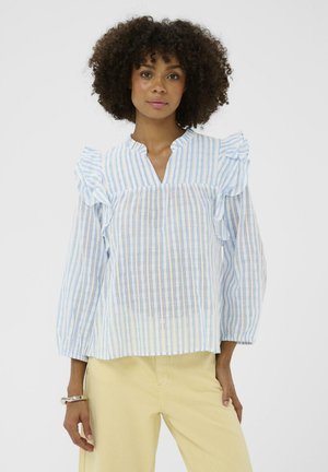 Sieviete ar cirtainiem matiem, valkājot zilas un baltas vertikālas svītras blūzi ar volāniem uz pleciem un gaiši dzeltenas bikses, stāvot pret balto fonu.