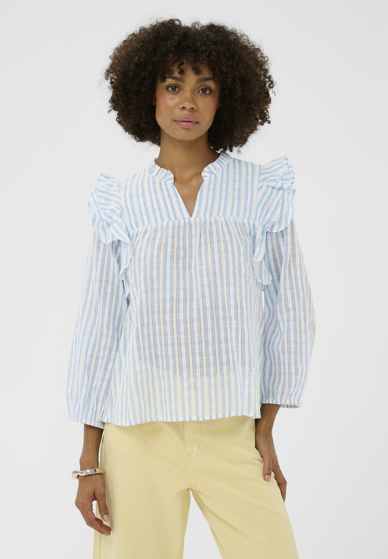 Femme aux cheveux bouclés portant un chemisier à rayures verticales bleues et blanches avec des volants aux épaules et un pantalon jaune clair, debout devant un fond blanc.