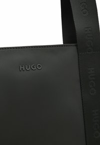 Black fabric tote bag with stitched edges, embossed "HUGO" logo on front, and matching black strap with repeated "HUGO" text.