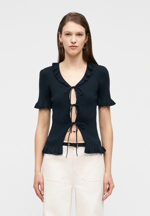 Frau trägt ein schwarzes, kurzärmeliges Strickoberteil mit drei Binderverschlüssen vorne und weiße, hoch taillierte Hosen mit einem schwarzen Gürtel.