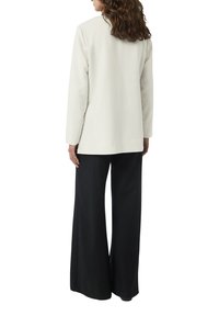 Helles beige Langarmoberteil mit geradem Schnitt, kombiniert mit weiten schwarzen Hosen. Glatter Stoff, minimalistisches Design, keine sichtbaren Muster oder Akzente.
