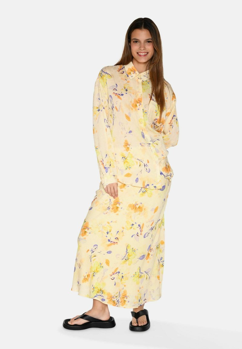 Blusa a maniche lunghe floreale giallo chiaro e gonna maxi coordinata con motivi blu e arancioni. Infradito nere completano l'outfit.