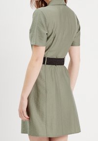 Robe à manches courtes en tissu vert olive clair, avec une taille cintrée et une ceinture élastique noire, et un col. Texture lisse.