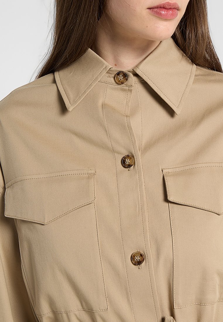 Beżowa koszula zapinana na guziki z kołnierzykiem, dwoma kieszeniami na piersi oraz fakturowanym materiałem. Posiada brązowe detale guzikowe.