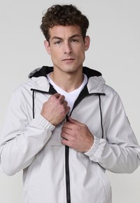 Giacca a vento leggera, grigia con accenti neri, polsini elasticizzati e cappuccio regolabile. Presenta una zip frontale e una tasca sul petto.