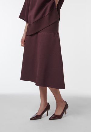 Femme portant une jupe midi bordeaux assortie à des escarpins pointus, debout devant un fond clair uni.