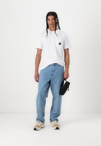Tricou alb cu mânecă scurtă și buzunar pe față, asortat cu blugi lejeri de un albastru deschis și adidași bej, ținând un pachet mic negru.