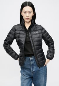 Melnā pūkainā jaka ar augstu apkakli un rāvējslēdzēju, ar izšūtu faktūru un EA7 logotipu uz krūtīm. Kombinēta ar melnu polo kreklu un zilām džinsa biksēm.