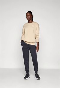 Beige genser med rund hals og hvitt logo, kombinert med mørkegrå joggebukser med matchende logo og elastiske mansjetter. Svarte joggesko.