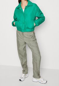 Person, der eine grüne Steppjacke, graue gerade Hosen und weiße Sneaker trägt, steht mit den Händen in den Jackentaschen vor einem schlichten Hintergrund.