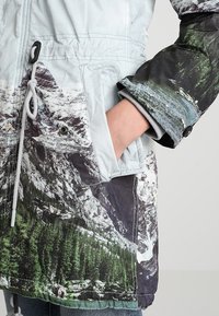 Monivärinen parkatakki, jossa on vuoristomaisemakuosi, säädettävä kiristysnyöri vyötäröllä ja sivutaskut. Valmistettu kevyestä, sileäpintaisesta kankaasta.