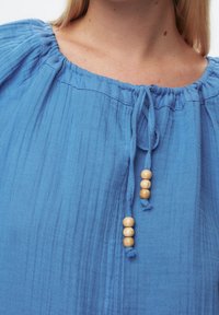 Blusa di cotone blu con una finitura testurizzata. Presenta un collo arricciato con lacci e dettagli in perline di legno. Vista ravvicinata del collo e del tessuto.