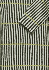 Gros plan d'un tissu en laine rayé verticalement noir et blanc avec de fines lignes horizontales jaunes et un détail de manche pliée.