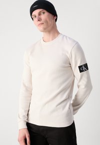 Svijetlosmeđa košulja dugih rukava s teksturiranim uzorkom i rebrastim manžetama. Ima crnu logotipsku oznaku na lijevoj ruci. Uparena s crnim hlačama.