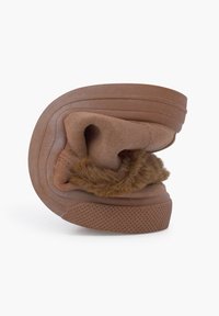 Zapatilla marrón con una suela de goma texturizada, parte superior de tela suave y forro de pelo sintético. Diseño enrollado para un ajuste flexible, que muestra un material suave y cálido.
