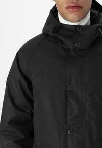 Σκοτεινό πράσινο αδιάβροχο μπουφάν με ψηλό γιακά, μαύρα κουμπιά και λεία υφή. Διαθέτει κλείσιμο με κουμπί μπροστά και μινιμαλιστικό σχέδιο.
