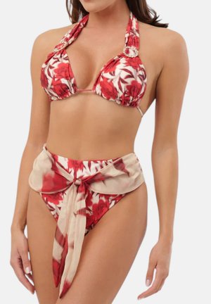 Donna che indossa un bikini a fiori rosso e bianco con top halter, slip a vita alta abbinato e una fascia beige e rossa a fiori annodata.