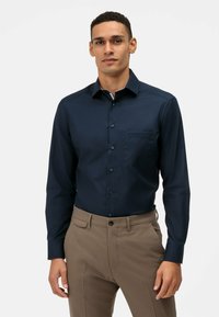 Marineblaues Langarmhemd mit Button-Down-Kragen, einer Brusttasche und dunklen Knöpfen. Wird mit beigen Hosen aus glattem Stoff getragen.