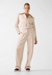 Veste beige à fermeture éclair avec col, associée à un pantalon à jambes larges de la même couleur. Tissu doux, coutures minimales, et poches latérales.