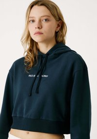 Mladá žena so svetlými vlasmi, oblečená v tmavej skrátenej mikine s nápisom "RED BULL RACING", stojaca na jednoduchom svetlom pozadí.