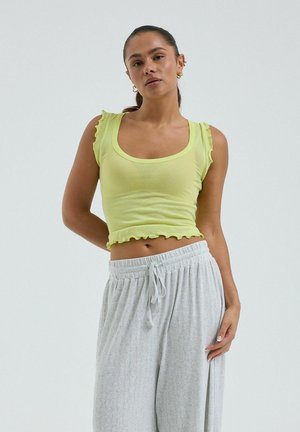 Femme debout, main derrière le dos, portant un haut sans manches vert citron avec des bords froncés et un pantalon gris clair à cordon de serrage.