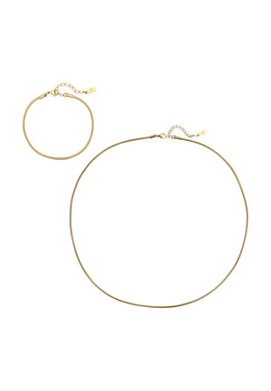 Männerhals mit kurzem Bart, der eine dünne goldene Halskette über einem weißen, gerippten ärmellosen Hemd trägt.