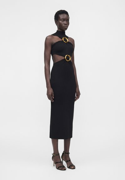 Ψηλή γυναίκα με αμάνικο μαύρο midi φόρεμα με ανοίγματα και χρυσούς κρίκους, σε συνδυασμό με μαύρα σανδάλια με ψηλό τακούνι.