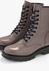 Paire de bottes de combat taupe brillantes avec des lacets noirs épais et des semelles en caoutchouc noir épaisses sur un fond clair.