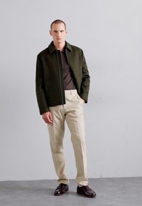 Olive green zip-up jacket, brown shirt, beige pants, brown leather shoes. Minimalist design with structured shape, smooth texture.