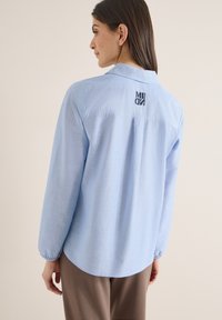 Femme aux cheveux bruns raides portant une chemise bleu clair à manches longues avec un logo brodé foncé sur le haut du dos, debout de profil.