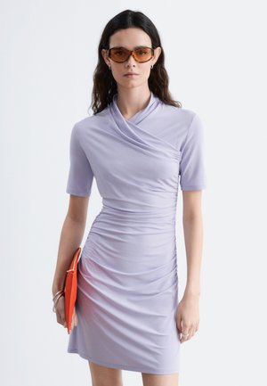 Femme aux longs cheveux foncés porte une robe lilas froncée, des lunettes de soleil teintées marron, des boucles d'oreilles anneaux argentées, des bracelets, et tient une pochette orange vif.