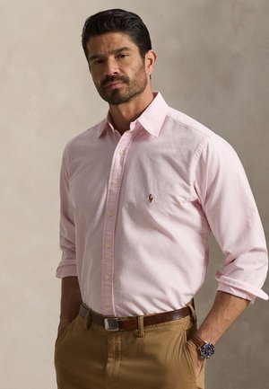 Uomo con la barba che indossa una camicia rosa chiaro con bottoni, pantaloni marroni e orologio da polso, in piedi con le mani in tasca davanti a uno sfondo uniforme.