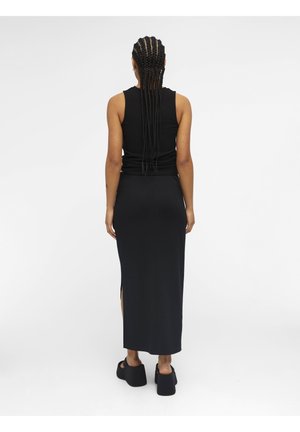 Femme aux longues tresses portant un top noir sans manches, une longue jupe noire fendue sur le côté, et des sandales à plateforme noires, debout de dos.