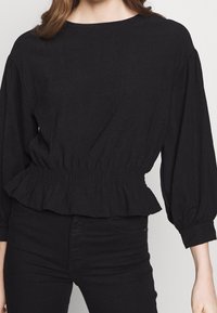 Top nero a maniche lunghe con scollatura rotonda, vita arricciata e maniche a sbuffo. Tessuto strutturato con una leggera lucentezza, aderente in vita.