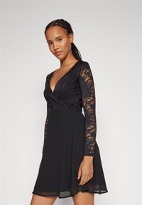 Μαύρο φόρεμα με σχέδιο περιτυλίγματος, διαφάνειες από δαντέλα με μακριά μανίκια και φουσκωτό κάτω μέρος. Μαλακό ύφασμα με περίπλοκα μοτίβα δαντέλας στα μανίκια.