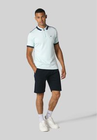 Gai zils polo krekls ar tumšām svītrām apkaklē, apģērbts kopā ar tumši ziliem šortiem. Baltas kedas un zeķes noslēdz apģērbu. Neitrāla fona.