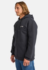 Navy fleece zip-up hoodie with drawstrings, front pockets, and embroidered logo. Soft texture with ribbed cuffs and hem. Casual fit.