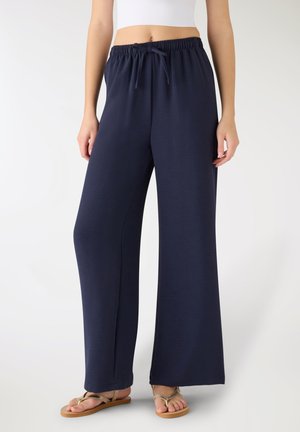 Donna che indossa pantaloni ampi blu navy con vita a coulisse e sandali beige, su uno sfondo chiaro e semplice.