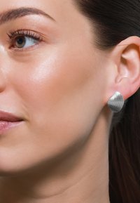 Orecchino a bottone in argento, con una texture a onde, di forma rotonda, fissato all'orecchio, che mette in risalto una superficie riflettente contro il tono della pelle.