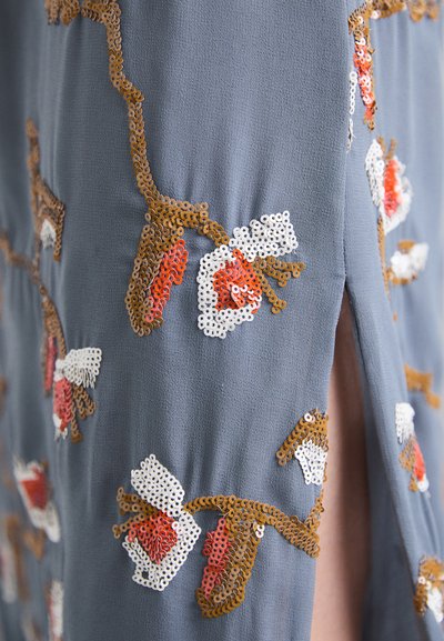 Αμπιγιέ φόρεμα με μπλε ύφασμα που διαθέτει λουλουδάτα μοτίβα από παγιέτες—καφέ κλαδιά, κόκκινα λουλούδια και λευκές λεπτομέρειες; λεπτομέρεια με πλευρικό σκίσιμο.