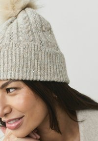 Gehaakte beige muts met een geribd ontwerp en een textuur van kabels, versierd met kleine pailletten en een bontige pompom bovenop.