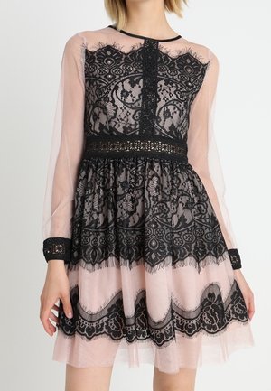 Robe longue noire en dentelle transparente à manches longues avec des motifs floraux, taille ajustée et doublure couleur chair, portée par une personne debout aux bras détendus.