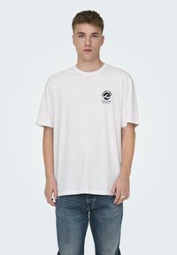 Weißes Baumwoll-T-Shirt mit kurzen Ärmeln, das ein kleines schwarzes Wellenlogo auf der linken Brust zeigt. Getragen mit blauen Jeans.