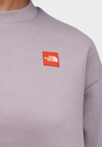 Μωβ φούτερ με ψεύτικο λαιμό και ριμπ υφή, με κόκκινο τετράγωνο λογότυπο με λευκό κείμενο από την The North Face.