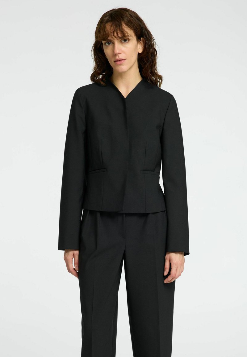 Frau mit schulterlangem braunem Haar, die ein tailliertes schwarzes Blazer und passende schwarze Hose trägt, steht vor einem schlichten Hintergrund.