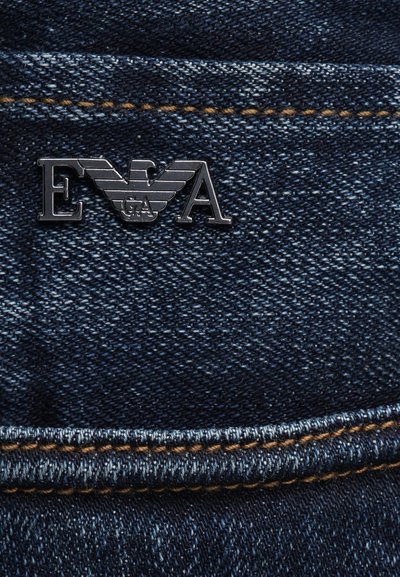 Mørkeblå denimstoff med oransje søm og en sølvfarget metalllogo med bokstavene E, A, og et vinget emblem med G A.