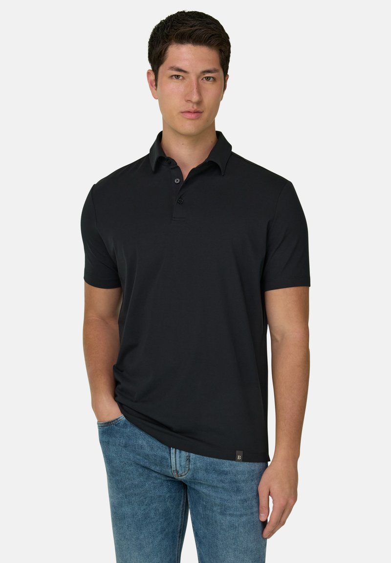 Jeune homme aux cheveux courts et foncés, portant un polo noir et un jean bleu, debout avec une main dans la poche.